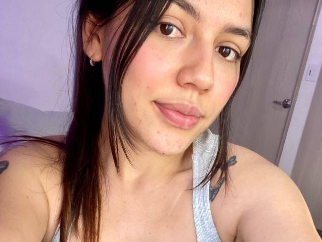 SophiaMarx with brown eyes, light skin, black hair, small nose, pink lips, wearing a gray tank top, showing tattoos on her shoulders, slight acne, and a subtle smile.