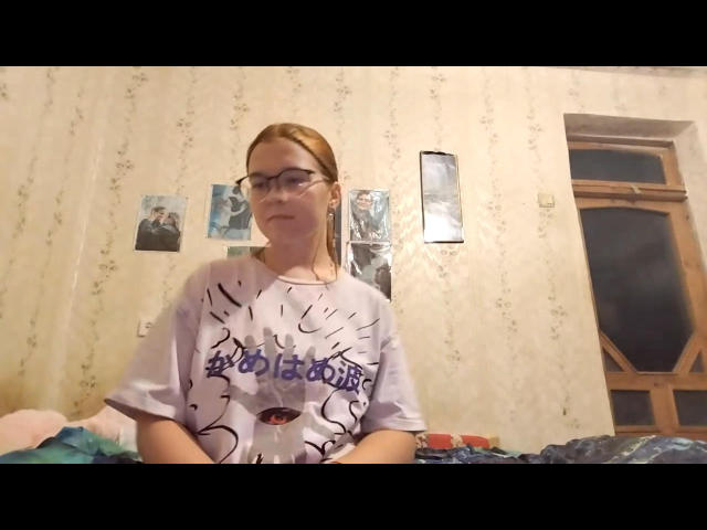 DrucillaSchab mit Brille, hellbraunes Haar im Pferdeschwanz, trägt ein violettes T-Shirt mit einem roten Auge und schwarzen Linien und sitzt auf einem Bett mit einer blauen Decke..
