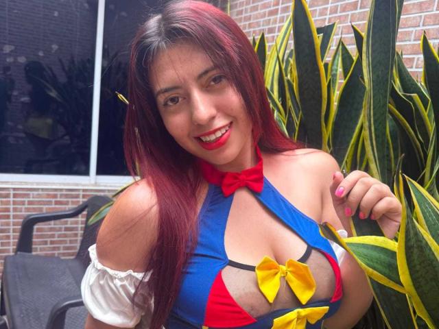 BrendaaPierce with red hair, smiling, wearing a blue, red, and yellow halter top that shows her tits, white puffed sleeves, and red bow, standing near green and yellow plants.