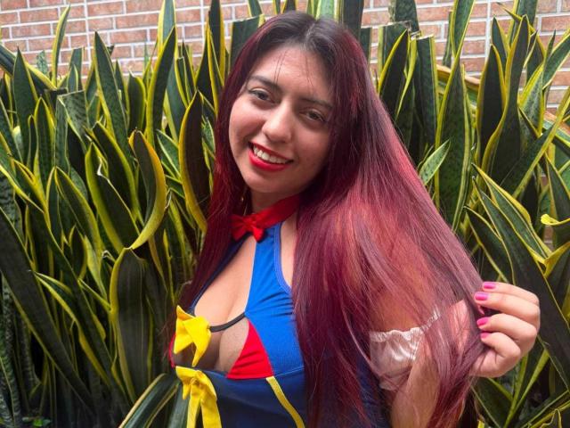 BrendaaPierce with long red hair, smiling, wearing a blue, red, and yellow outfit with a keyhole cutout showing her boobs, standing in front of green and yellow striped plants.