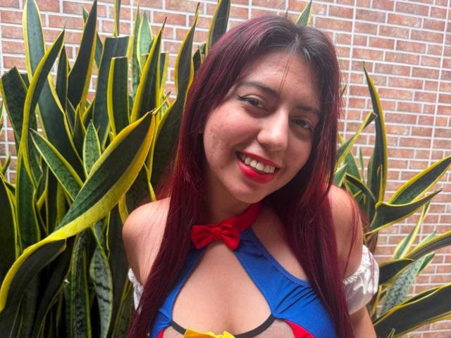 BrendaaPierce with red lipstick, smiling, has long dark red hair, wearing a blue and red costume with a red bow, showing her tits, standing in front of green snake plants.