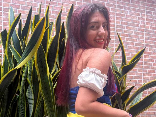 BrendaaPierce with long red hair, smiling, wearing an off-shoulder white and blue top, standing beside tall green and yellow snake plants, brick wall in background.