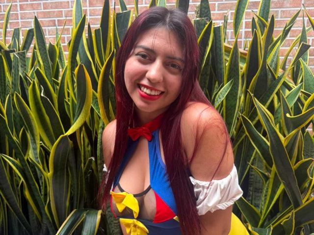 BrendaaPierce with red lipstick, smiling, long red hair, wearing blue and yellow costume with off-shoulder white sleeves, standing in front of green and yellow striped leaves.