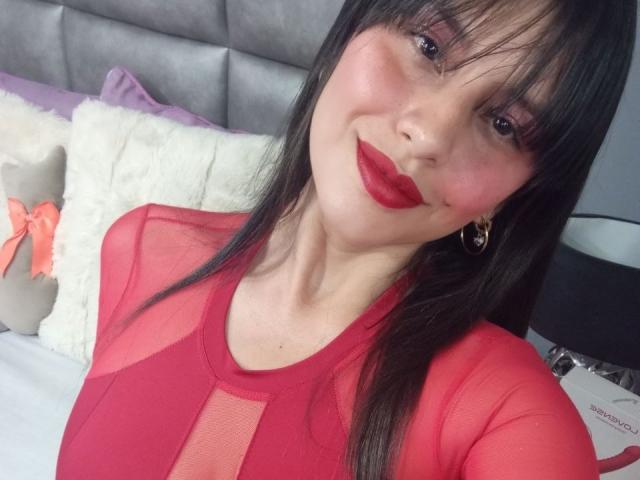 MadissonFontaine with black hair, red lipstick, red sheer top, and gold earrings, smiling at the camera. Boobs visible through the top, pink nipples not seen.