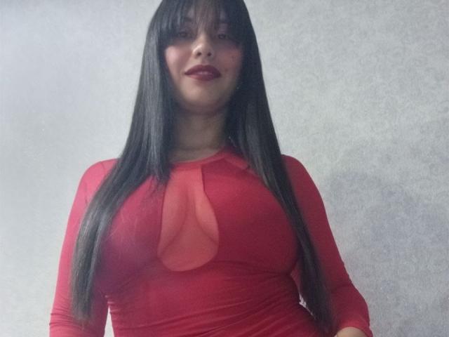 MadissonFontaine with long black hair, red lipstick, and a tight red dress with a keyhole cutout, showing her tits. She's standing against a plain, gray wall.