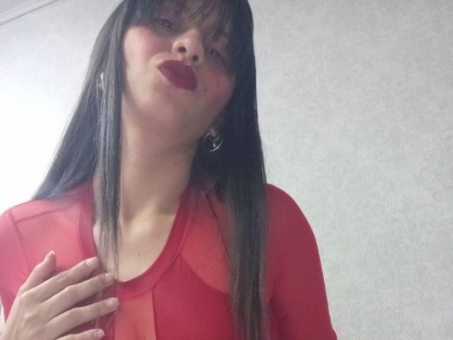 MadissonFontaine with long brown hair, red lipstick, and red sheer top, touching chest, wearing hoop earrings, looking up slightly, smiling. Boobs visible through translucent fabric.