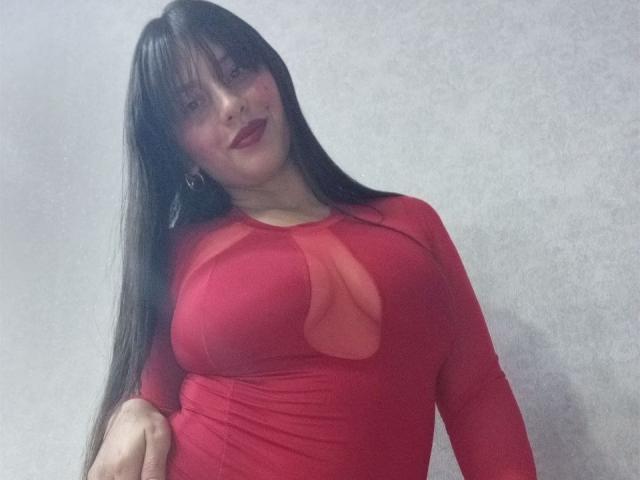 MadissonFontaine with long black hair, red dress, and big tits, smiling with dark lipstick, standing against a plain wall. Her hand rests on her hip.