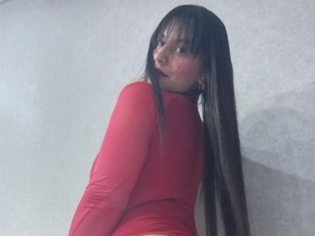 MadissonFontaine with long black hair, red long-sleeve dress, looking over shoulder, showing her ass, light skin, slight smile, gray wall background, subtle makeup.
