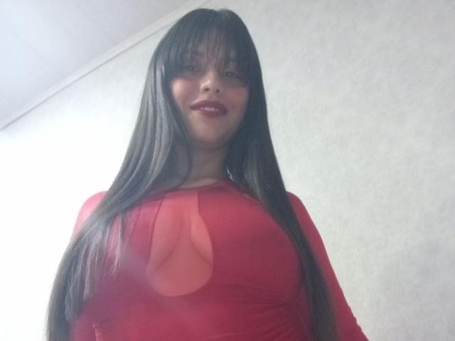 MadissonFontaine with long brown hair, red dress, and visible cleavage, smiling, red lipstick, standing against a white wall. Her tits are slightly exposed through the dress.