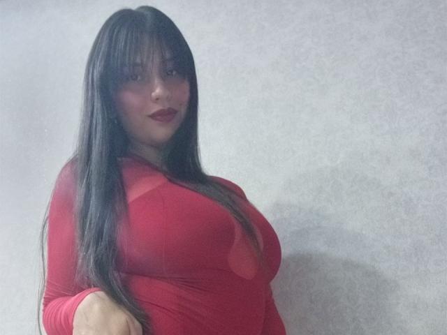 MadissonFontaine with long black hair, wearing a red, form-fitting dress that highlights her big tits. She's standing, looking at the camera with dark red lipstick.