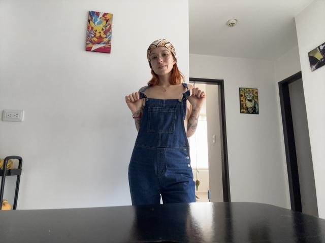 Sarah Rousen mit roten Haaren, die eine gemusterte Stirnband trägt, Jeanslatzhosen und Riemen hält, wobei Tattoos auf ihren Armen sichtbar sind, steht in einem Raum mit einem schwarzen Tisch im Vordergrund..