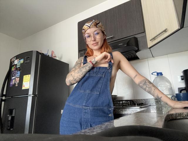 Sarah Rousen, gekleed in een denim overall, met een bril en bandana, staat in een keuken met tatoeages, haar kleine borsten vasthoudend, met één arm op het aanrecht, en kijkt naar de camera..