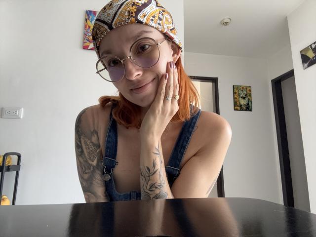 Sarah Rousen, com cabelo ruivo, usando um gorro estampado, óculos redondos e macacão de jeans, tocando sua bochecha, mostrando tatuagens, sentada em uma mesa preta, sorrindo suavemente..