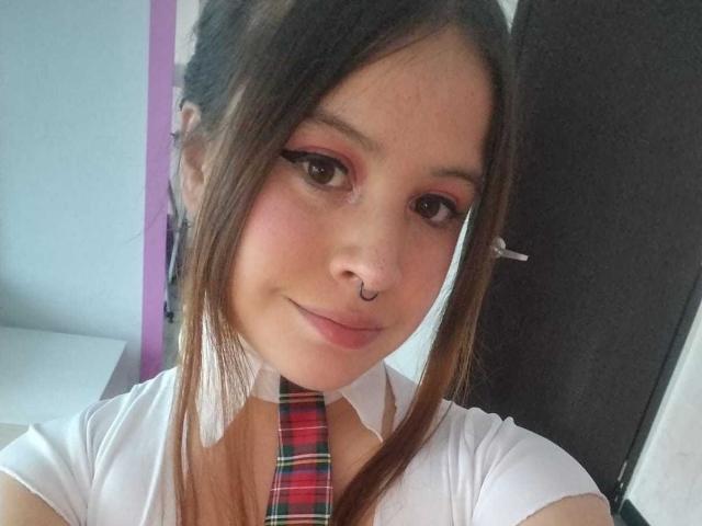 Melani Becker heeft bruine ogen, roze oogschaduw en een neusring. Ze draagt een witte blouse met een rode geruite das en heeft een lichte glimlach. Bruin haar, haarspeldjes..