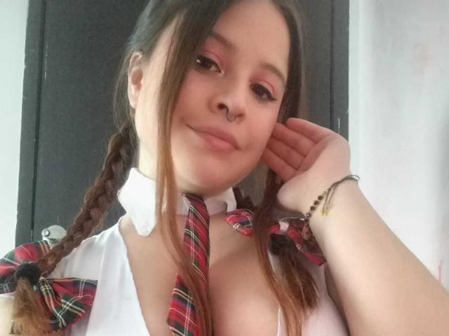 Melani Becker met gevlochten bruin haar, roze oogmake-up, neusring, wit shirt, geruit dasje en strik, lachend naar de camera, hand op haar wang, waarbij haar borsten zichtbaar zijn..