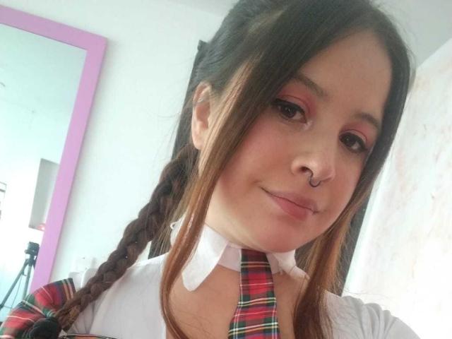 Melani Becker, met gevlochten bruin haar, roze oogschaduw, een neusring, een wit shirt en een geruit dasje, glimlacht zachtjes naar de camera. Borsten niet zichtbaar..
