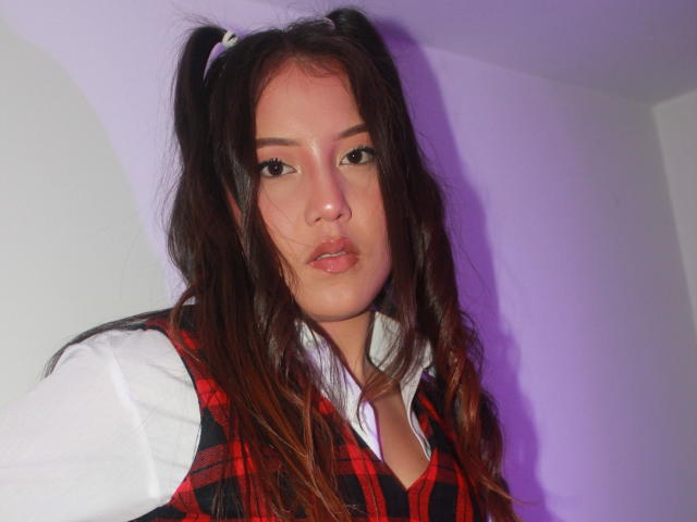 SweetTem, con le labbra piene, indossa un gilet a quadri rossi sopra una camicia bianca, ha lunghi capelli castani raccolti in due trecce, e un leggero ombretto viola sul viso..