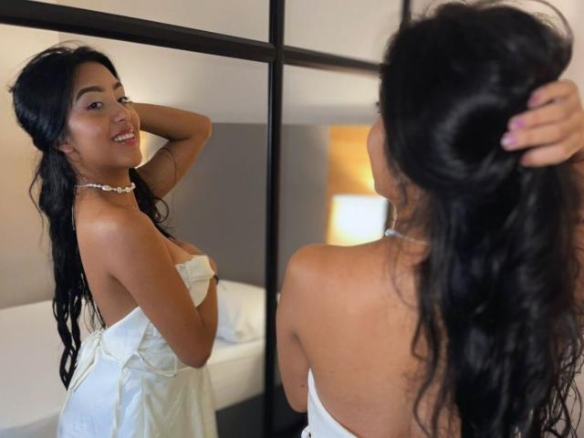 EmmaEstreilla with long black hair, smiling, wearing a white strapless dress, necklace, and holding her hair up, reflected in a mirror, showing her back and tits.