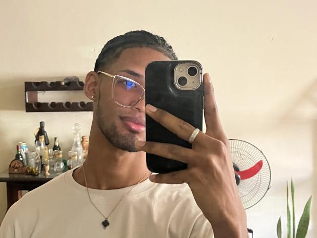 DanielNaugthyOne with glasses, holding a phone, white tee, ring, and necklace, taking a selfie, showing pink lips, and a slight beard, with braided hair, in a mirror.