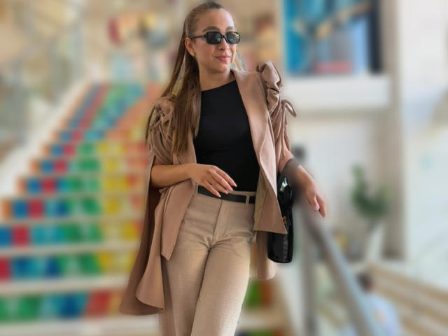 Arianna Blake, con cabello rubio, blusa negra, pantalones beige y chaqueta color arena, sosteniendo un bolso negro, caminando por unas escaleras coloridas, usando gafas de sol negras, ligeramente borroso..
