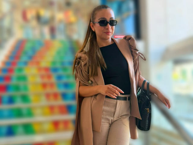 AriannaBlake with long brown hair, black sunglasses, tan coat, black top, white pants, black bag, standing, looking slightly to the right, holding coat shoulder.