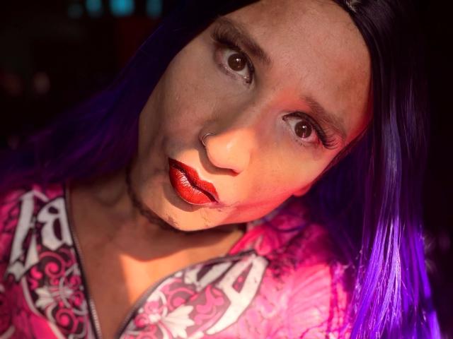 MaryXXL, con cabello morado, lápiz labial rojo, piercing en la nariz y labios rojos brillantes, vestida con una camisa rosa con estampado floral, bajo una luz dramática. Su rostro está ligeramente inclinado, con los ojos mirando hacia adelante..