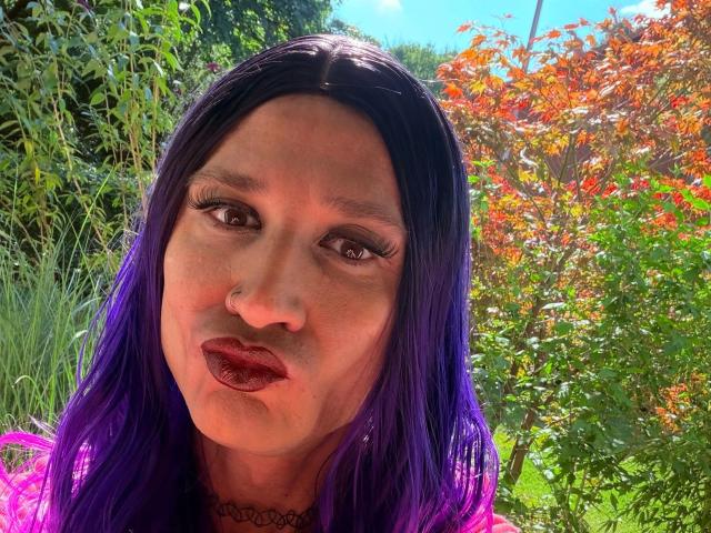 MaryXXL-hot with purple hair, dark lipstick, and a black choker, pouting in a sunny garden. Her makeup includes long, thick eyelashes. She's wearing a purple top.