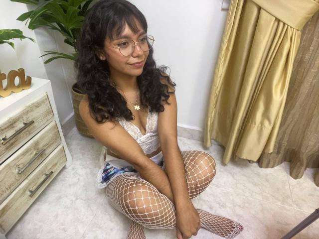 JuliaBennett, cute brown-skinned woman with curly black hair, small tits, wearing glasses, white lace top, blue plaid skirt, fishnet stockings, sitting on white tile floor, smiling.
