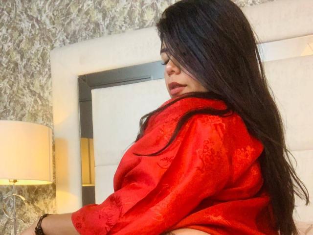 CatalinaReiyna in a red, silky robe with floral patterns, long black hair, side profile, looking down, sitting on a bed, next to a white lamp, soft lighting.