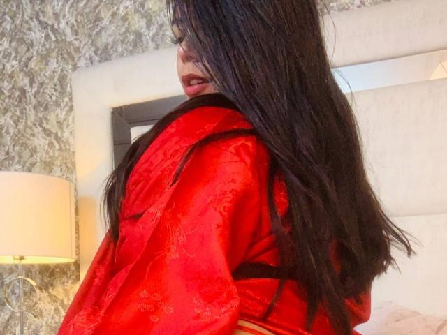 CatalinaReiyna with long black hair, red silk robe, and visible red lipstick, looking over her shoulder, showing her back and a hint of her ass.
