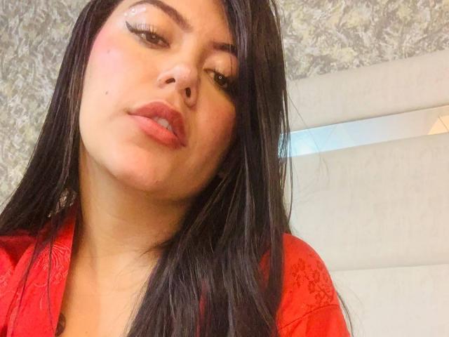 CatalinaReiyna with long black hair, red lips, and a slight pout, wearing a red dress, looking down with a subtle smirk, showing her smooth, medium-sized tits.