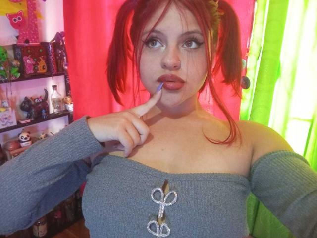 SophieHill with red pigtails, gray off-shoulder top, button down, finger in mouth, looking up, nose ring, blue lipstick, cute, with small tits, in a colorful room.