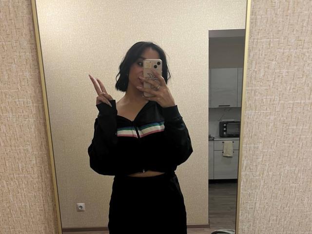 Charleen Oachs, com cabelo preto curto, vestindo uma blusa preta com listras coloridas de arco-íris e uma saia preta, fazendo o sinal de paz, segurando um celular em uma selfie no espelho..