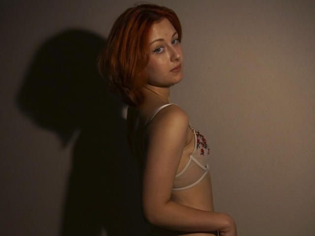 Masako Parkey, con cabello rojo, vistiendo un sujetador blanco transparente con bordados florales rojos, mirando por encima del hombro, sombra en la pared, iluminación suave, piel pálida, maquillaje sutil..