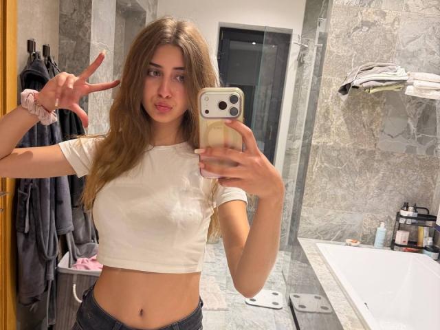 JameStueber with long brown hair, white crop top, showing her flat tits, making a peace sign, taking a mirror selfie in a bathroom with marble walls.