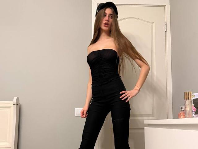 JameStueber, con lunghi capelli castani, indossa un top nero senza spalline che mette in mostra il seno, pantaloni neri, un cappello nero, con una mano appoggiata al fianco, in piedi in una stanza con una porta e un armadietto bianco..