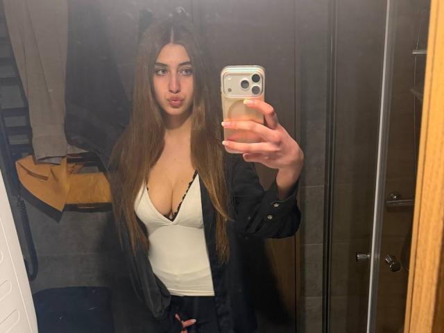 JameStueber with long brown hair, pouty lips, and large tits, wearing a white tank top and black shirt, taking a mirror selfie with a smartphone in a bathroom.