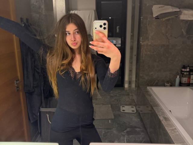 JameStueber, con lunghi capelli castani, indossa una maglia nera con maniche lunghe in pizzo e pantaloni neri, e sta scattando un selfie allo specchio con uno smartphone, con un'espressione leggermente accigliata..