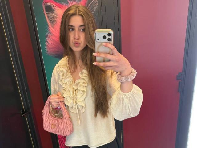 JameStueber, con cabello castaño largo, labios carnosos, blusa blanca con volantes, una goma para el pelo rosa, sosteniendo un bolso rosa, tomando una selfie con su iPhone, con una pared rosa de fondo..