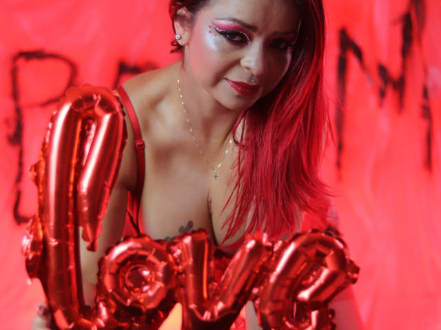CataleyaSweett with red hair, wearing a red bra, holding shiny red "love" balloons, smiling, makeup with pink eyeshadow, necklace, and tattoo visible on her chest.