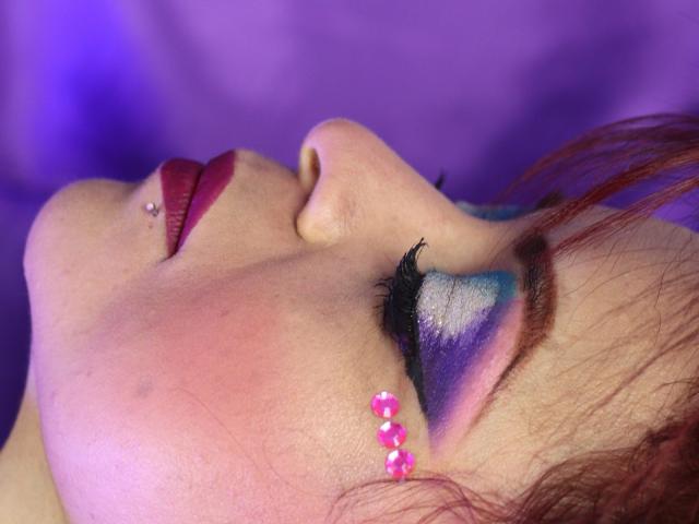 Le visage de CataleyaSweet, avec un maquillage oculaire violet et bleu, des perles roses sur la joue, du rouge à lèvres rouge, un piercing au nez et les yeux fermés, sur un fond violet..