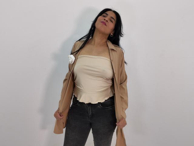 Rhouss with long black hair, tan jacket, white strapless top, and dark jeans, holding jacket sleeves, looking down with slightly parted pink lips, small white necklace.