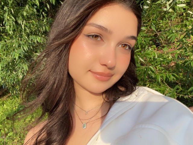 Sofia, avec ses cheveux ondulés brun moyen, ses yeux marron clair et un sourire discret, porte une chemise blanche qui laisse apparaître ses épaules, et une chaîne avec un petit pendentif..