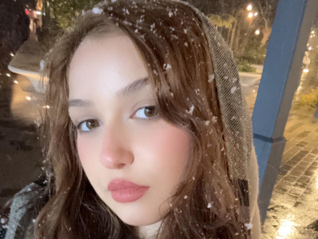 SofiaShow ha un viso carino con rossetto rosa, capelli castani ondulati e fiocchi di neve sulla testa. Indossa un cappuccio beige e guarda di lato..