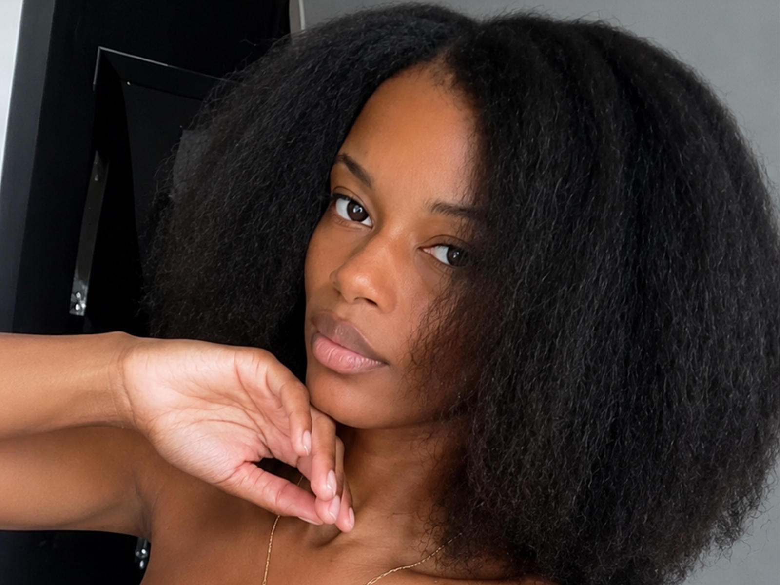 NicolCastillo, avec ses cheveux bruns et bouclés, ses yeux marron et son collier en or, se touche le menton avec une expression détendue, légèrement sérieuse. Sa peau lisse et brune est visible..