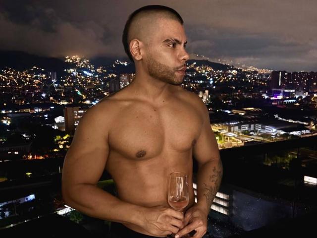 AdrianMontrell ist ohne Hemd, hat einen muskulösen Körperbau, hält ein Glas rosa Wein und blickt zur Seite. Er ist kahlgeschoren und hat Tattoos am linken Arm..