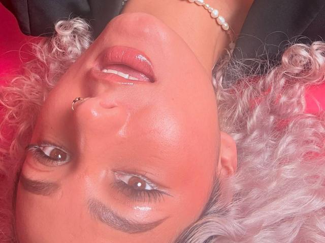 ElenaSaenz's shiny pink lips are slightly open, showing her white teeth. She has curly silver hair, a nose ring, and a pearl bracelet on her wrist. Red background.