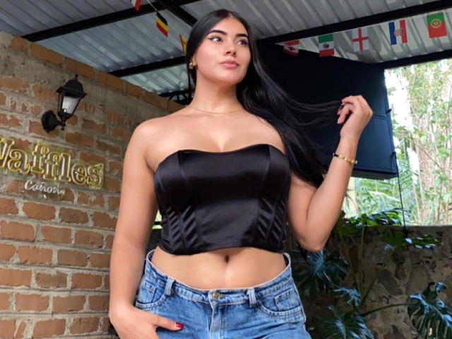 AlaraKey with long black hair, wearing a black strapless top, showing her tits, and blue jeans, holding her hair, standing in front of a brick wall.