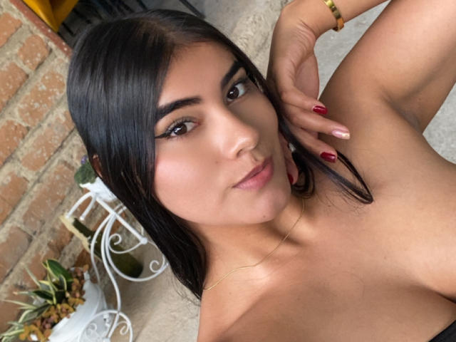 AlaraKey with dark hair, brown eyes, and red nails, looking at the camera with a slight smile, showing her bare shoulders and a gold bracelet.