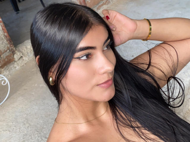 AlaraKey with long black hair, tan skin, and subtle makeup, showing off her tits by raising an arm, wearing a gold bracelet and small earrings.